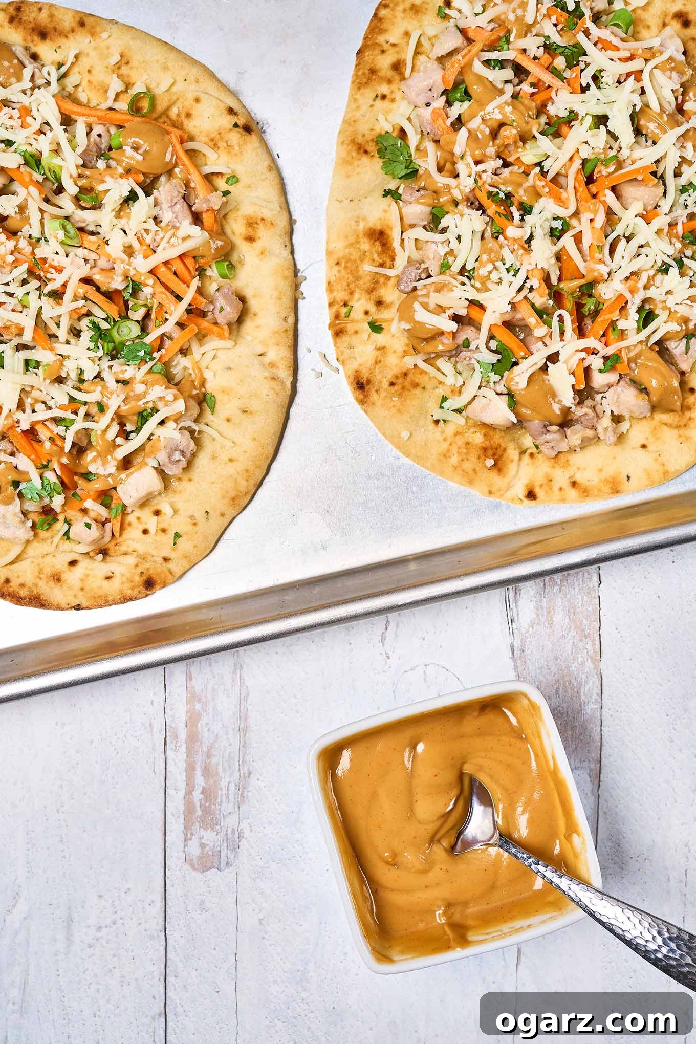 Spicy Thai Chicken Flatbread 5 A prep photo showing two chicken flatbreads assembled with toppings and ready to be baked atop a baking sheet lined with parchment paper, with a bowl of peanut sauce on the side.
