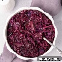 An overhead view of a serving bowl of braised red cabbage with a spoon in it, on top of a linen napkin with white ceramic salt and pepper shakers behind it and to the left.