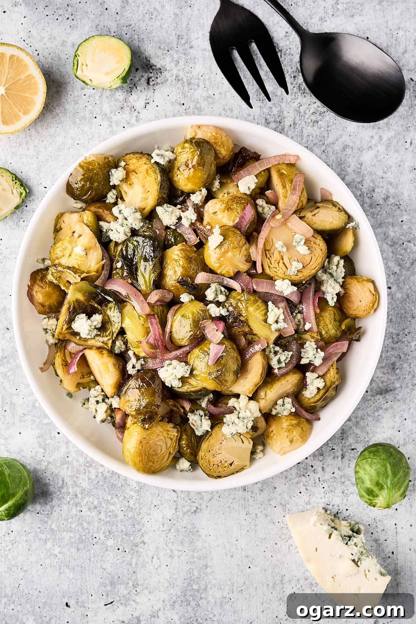 Bold Blue Brussels Sprouts 2 A bowl of roasted Brussels sprouts, red onion, and blue cheese on a gray surface, surrounded by utensils, scattered Brussels sprouts, lemon, and blue cheese.