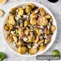 A bowl of roasted Brussels sprouts, red onion, and blue cheese on a gray surface, surrounded by utensils, scattered Brussels sprouts, lemon, and blue cheese.