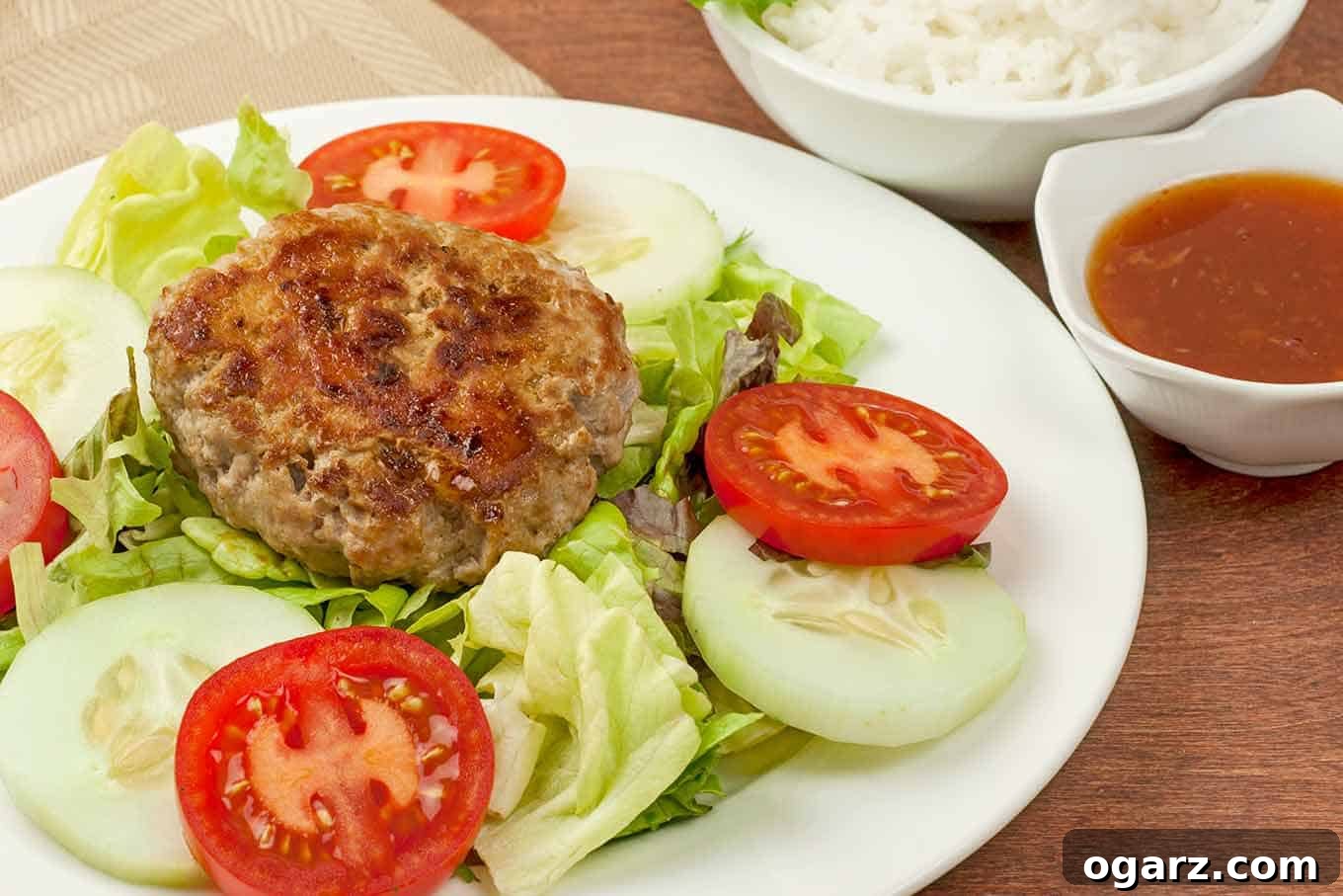 Bed of lettuce, tomatoes, and cucumbers topped with a pork patty, with rice and dipping sauce on the side.