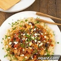 A dinner plate with boneless chicken breasts, topped with a light sauce of tomatoes, Kalamata olives and feta cheese served over a bed of riced cauliflower, extra plates and napkins in the background.
