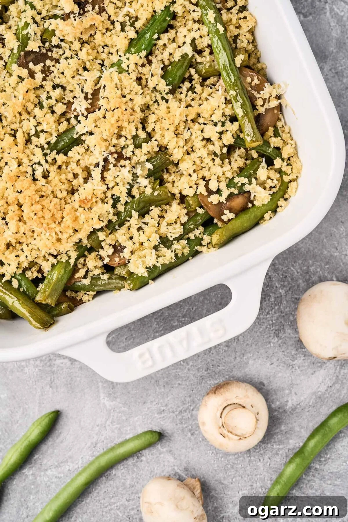 Golden Crumb Green Bean and Mushroom Medley 8 A beautiful baking dish filled with green beans and mushrooms, crowned with a golden, crispy crumb topping, presented on a gray surface with fresh, raw mushrooms and green beans beside it, showcasing the ingredients and final dish.