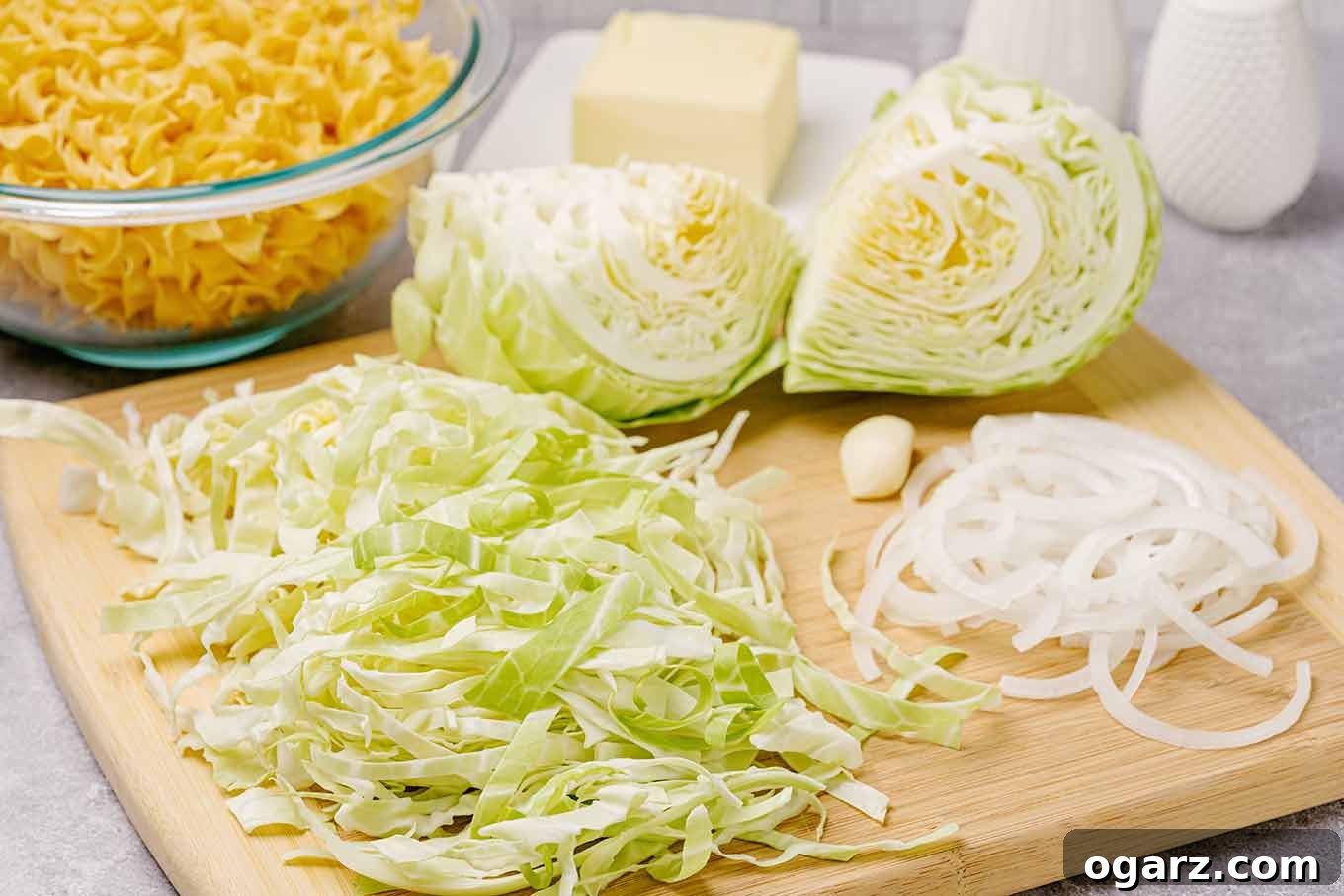 A vibrant display of fresh ingredients for Hungarian Cabbage and Noodles, including a mound of thinly-sliced green cabbage, cabbage wedges, meticulously sliced onion, garlic cloves, alongside a package of wide egg noodles and a stick of unsalted butter.