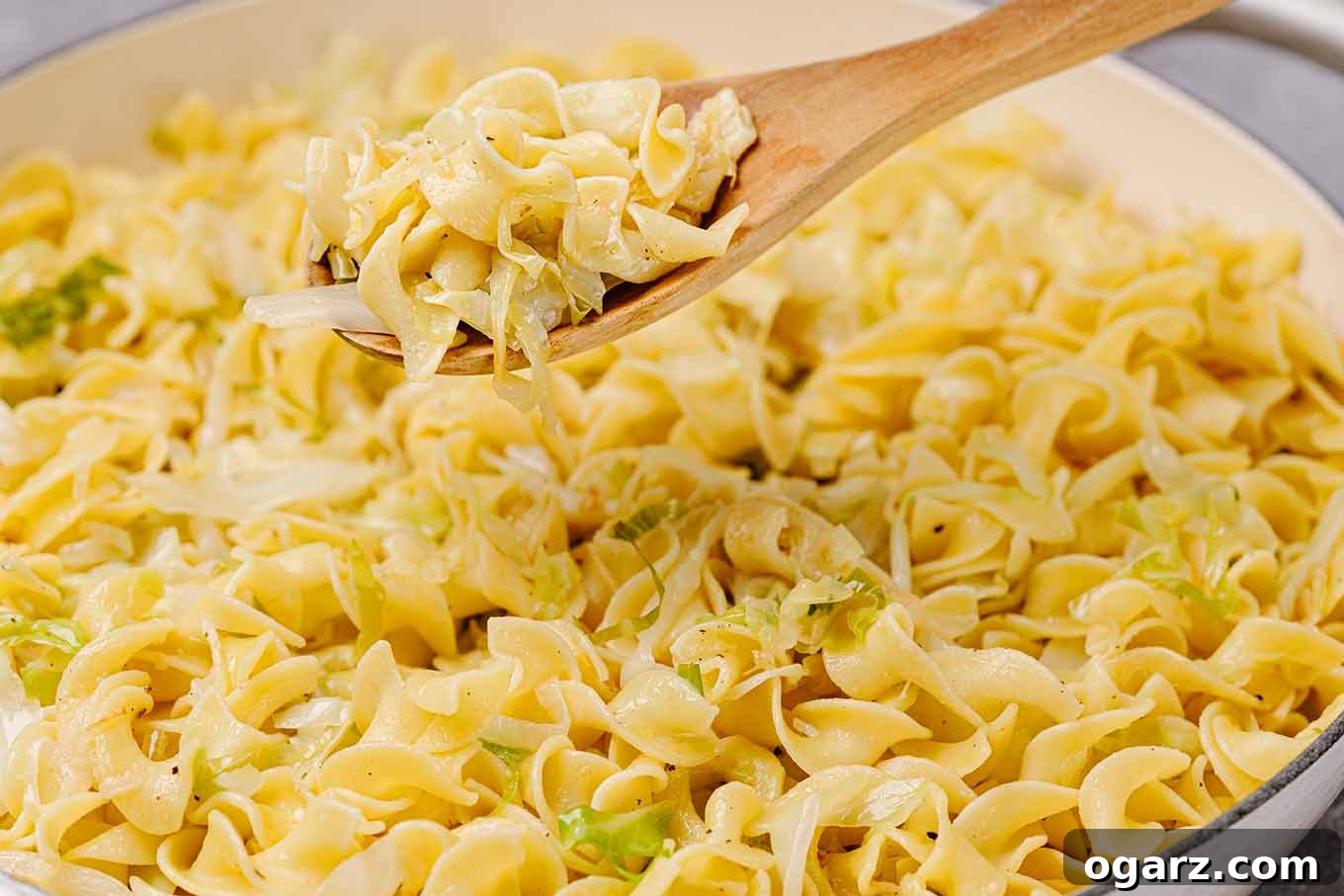A detailed close-up shot of a cast iron pan brimming with perfectly sautéed cabbage and golden egg noodles. The focus is on a wooden spoon lifting a generous portion, highlighting the texture and deliciousness of the dish.