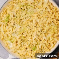 An enameled cast iron pan filled with a combination of egg noodles and sliced cabbage sautéed in butter and a wooden spoon on the counter.