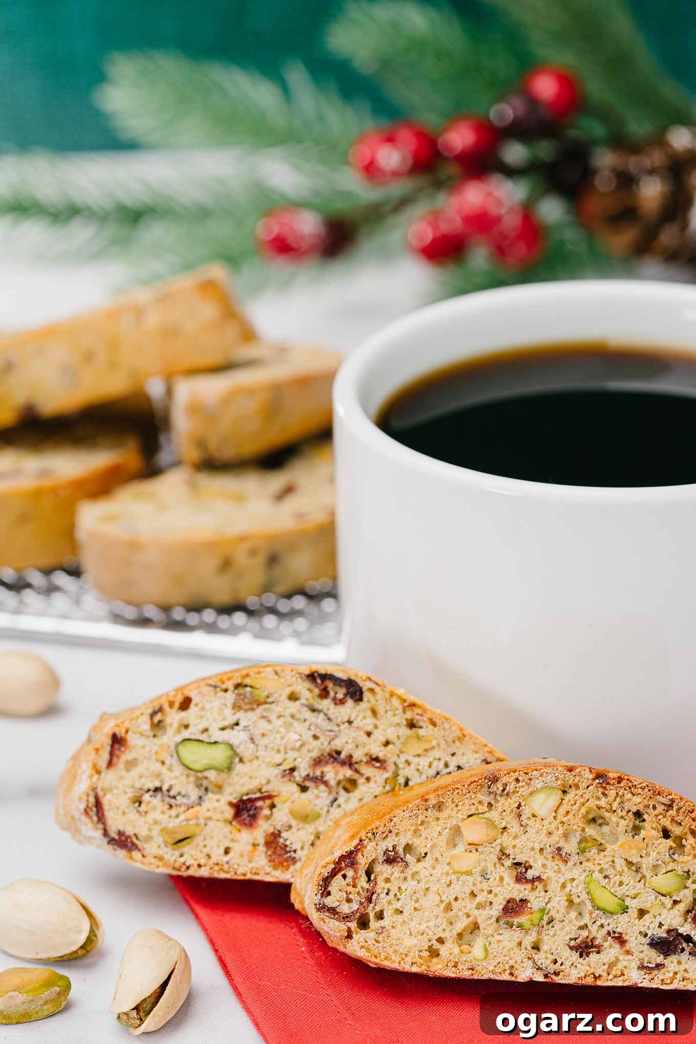Pistachio & Cranberry Biscotti 2 Two cranberry pistachio biscotti on a red napkin next to a cup of black coffee with more biscotti in the background, inviting a warm, comforting experience.
