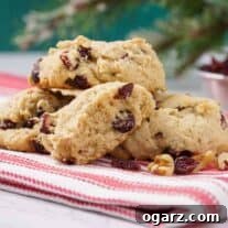 Cranberry walnut cookies on a folded red and white towel with dried cranberries and nuts scattered in front, a dish of dried cranberries in the background.