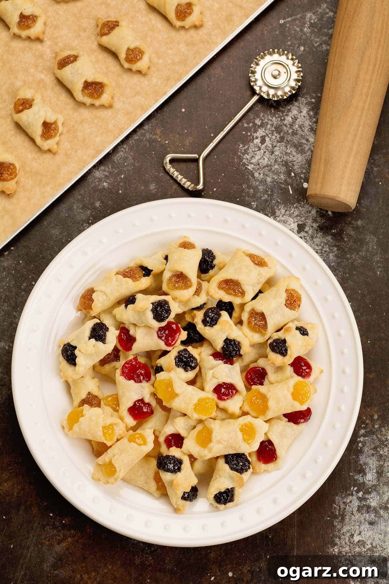 Our Family's Treasured Hungarian Kiffle Recipe 2 A white plate of assorted flavors of Hungarian kiffles with a cookie sheet, pastry wheel, and rolling pin in the background. Traditional Hungarian cream cheese pastry cookies.