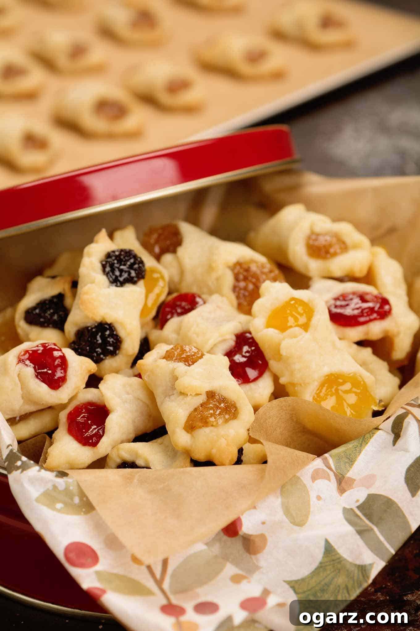 Our Family's Treasured Hungarian Kiffle Recipe 4 A red Christmas gift tin filled with apricot, almond, poppy seed, and raspberry Hungarian kiffles, with a baking sheet of more freshly baked kiffles in the background. A delightful holiday treat.