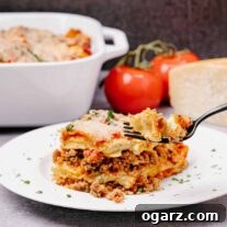 A single serving of sausage lasagna with a forkful being lifted up, the baking dish, fresh tomatoes, and a wedge of cheese in the background.