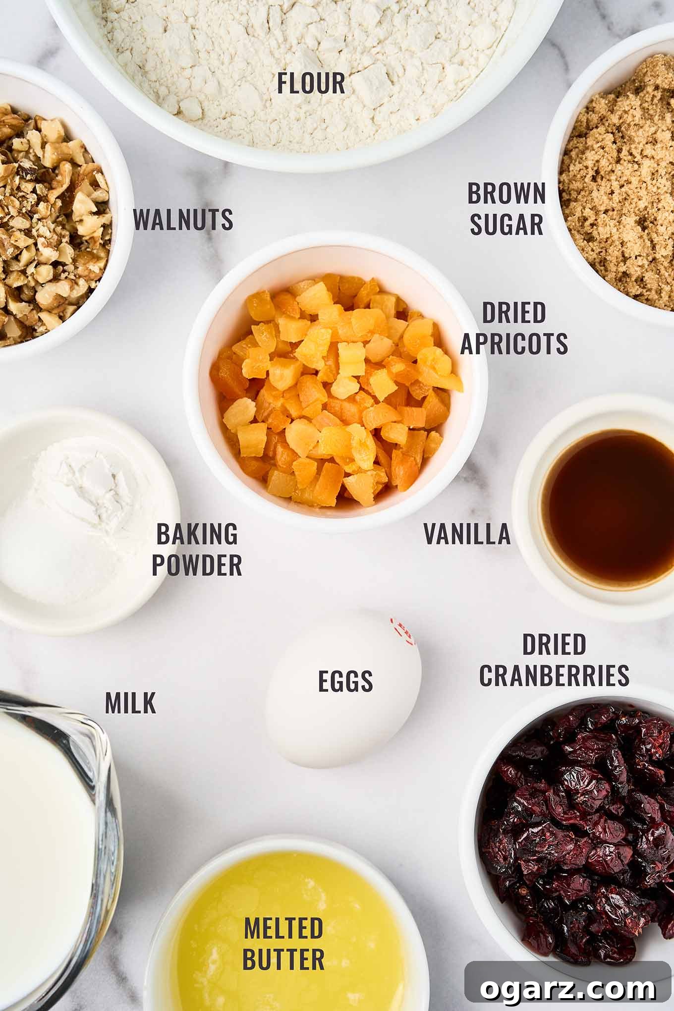 Golden Apricot Cranberry Loaf 3 A labeled, overhead view of the ingredients for apricot quick bread, including flour, brown sugar, dried apricots and cranberries, walnuts, baking powder, milk, eggs, vanilla, and melted butter.