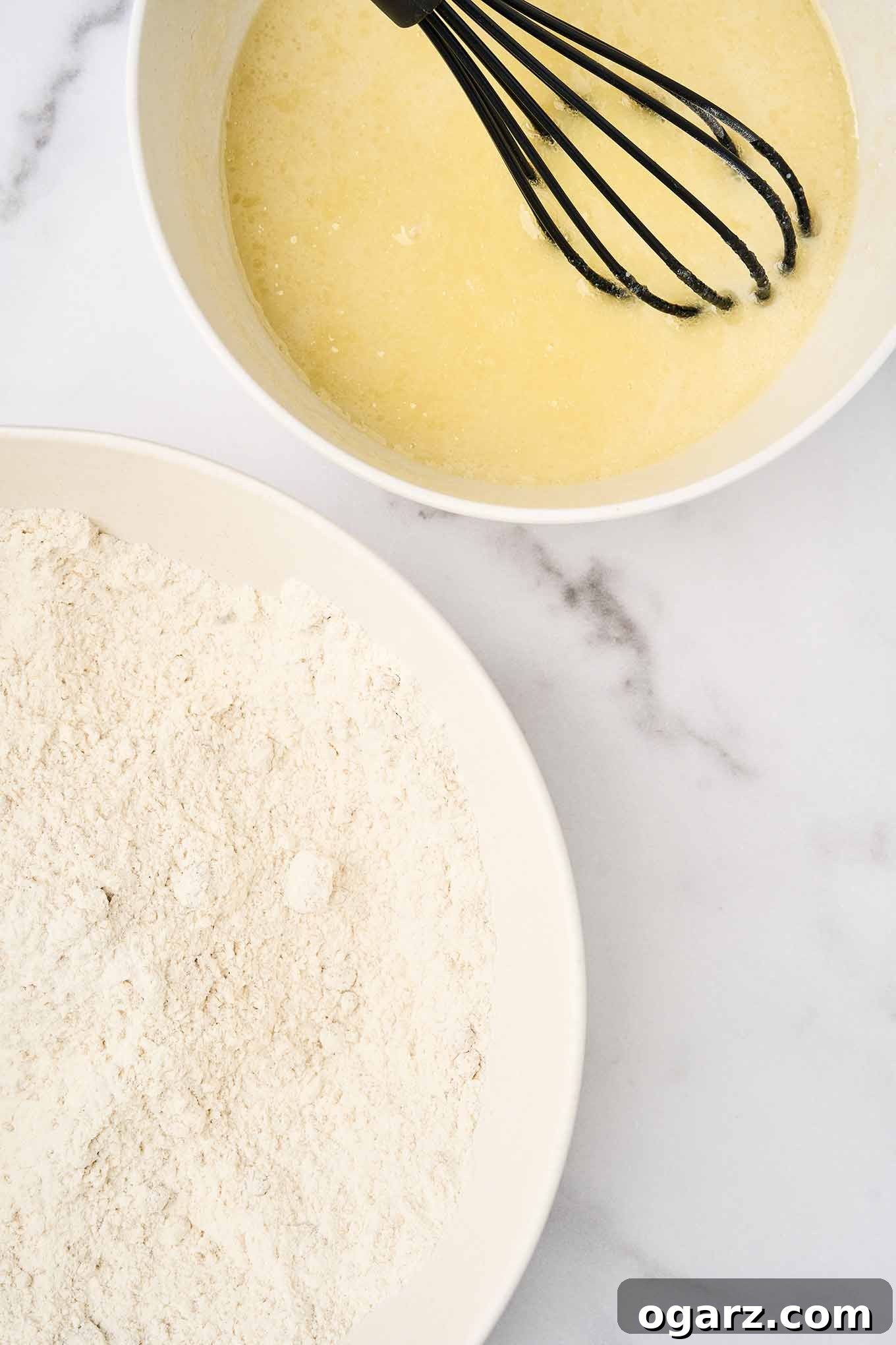 Golden Apricot Cranberry Loaf 4 Two mixing bowls side by side: one with flour and dry ingredients, the other with whisked wet ingredients and a black whisk, set on a marble surface.