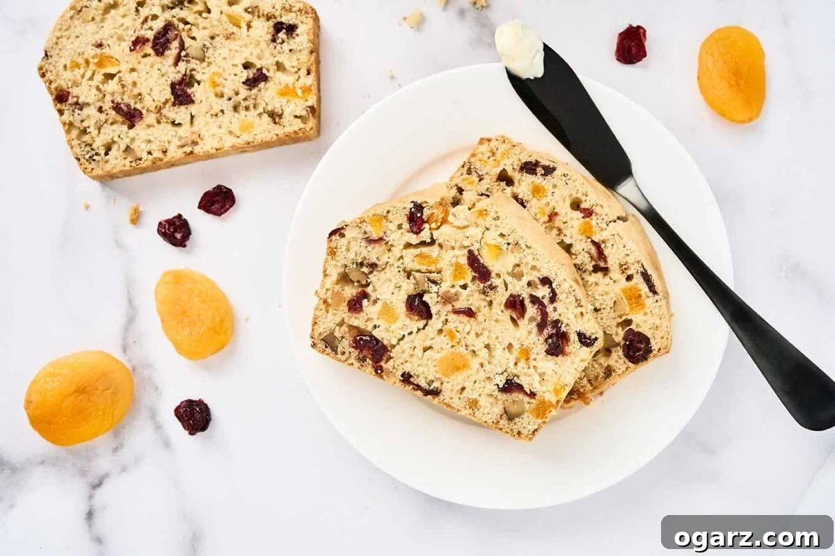 Golden Apricot Cranberry Loaf 6 Two slices of apricot cranberry quick bread on a white plate with a buttered knife, surrounded by scattered dried fruits on a marble surface.