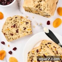 Sliced apricot bread with cranberries and walnuts on a white marble surface, surrounded by dried apricots, cranberries, and a buttered knife.