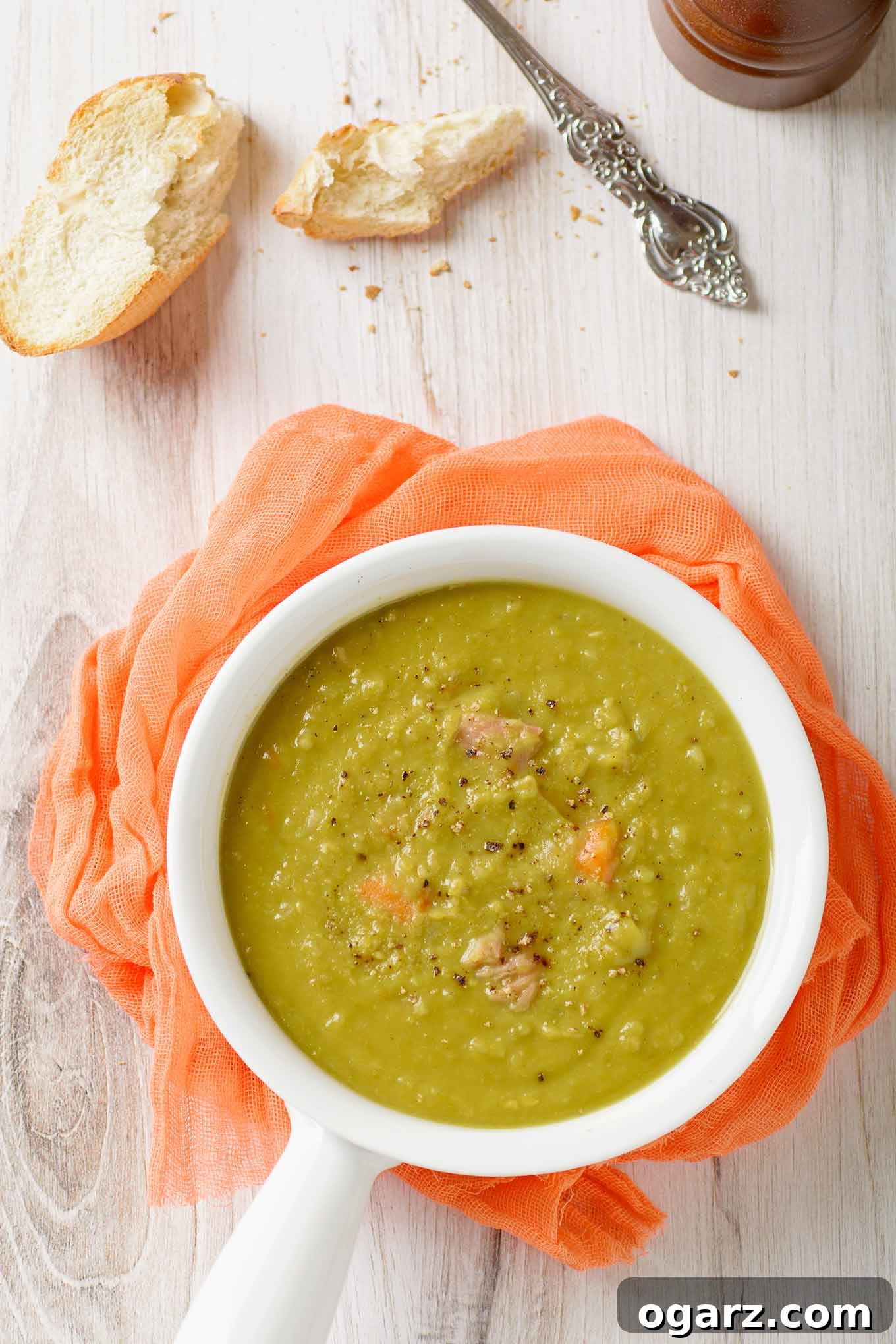 Simple Homestyle Split Pea Soup 2 A serving of split pea soup in a white bowl with a handle on a light wooden surface with crusty bread, soup spoon, and pepper mill on the side.