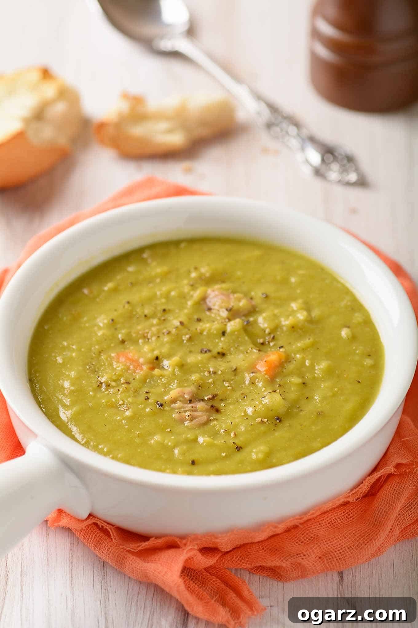Simple Homestyle Split Pea Soup 3 A serving of split pea soup in a white bowl with a handle on a light wooden surface with crusty bread, soup spoon, and pepper mill on the side.