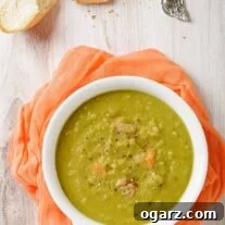 Simple Homestyle Split Pea Soup 4 A serving of split pea soup in a white bowl with a handle on a light wooden surface with crusty bread, soup spoon, and pepper mill on the side.