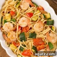 An overhead view of an oval casserole filled with sautéed zucchini and red bell pepper tossed in a light garlic sauce with linguine and shrimp.