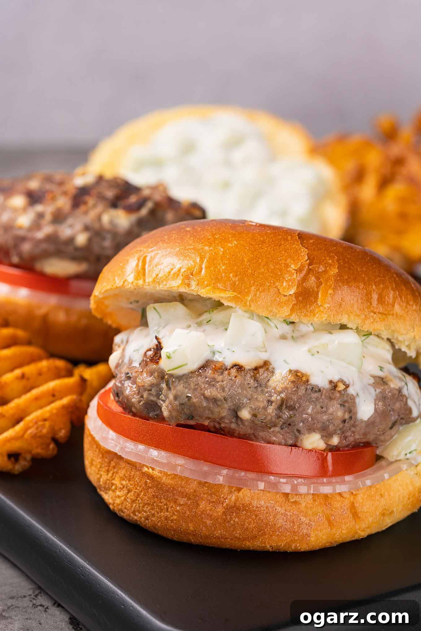 Grecian Lamb & Herb Burgers 2 Two grilled lamb burgers on brioche buns with sliced onion and tomato and a topping of tzatziki sauce, on a serving board with fries.