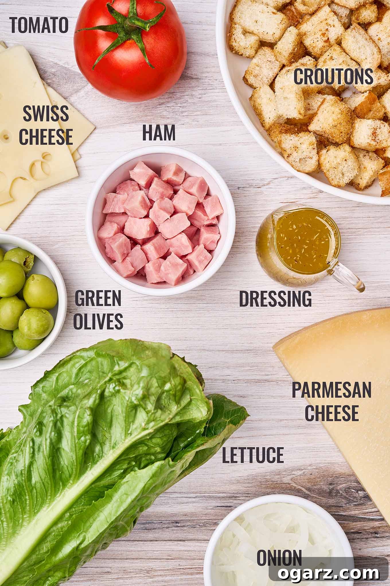 Columbia's Signature 1905 Salad 3 A labeled overhead view of 1905 salad ingredients; cubed ham, Swiss cheese, lettuce, green olives, croutons, a tomato, Parmesan cheese, and dressing.