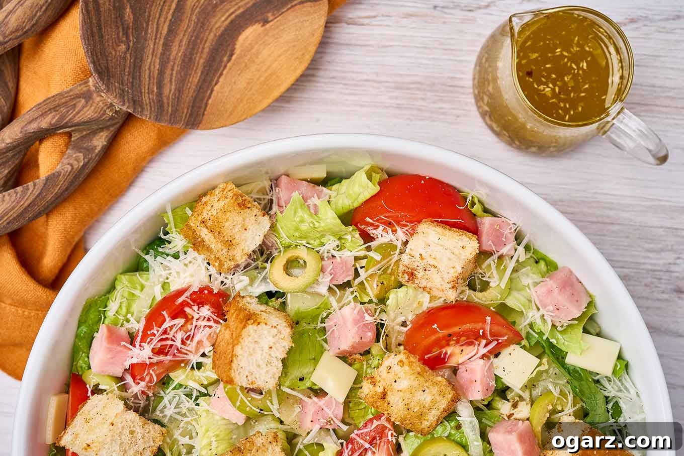 Columbia's Signature 1905 Salad 5 A serving of 1905 salad in a white bowl on a wooden background next to a clear glass pitcher of dressing and a linen napkin and wood salad servers on top.