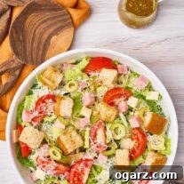 A dinner-sized serving of 1905 salad in a white bowl with croutons, grated Parmesan, olives and cubes of ham; wooden salad servers to the side.