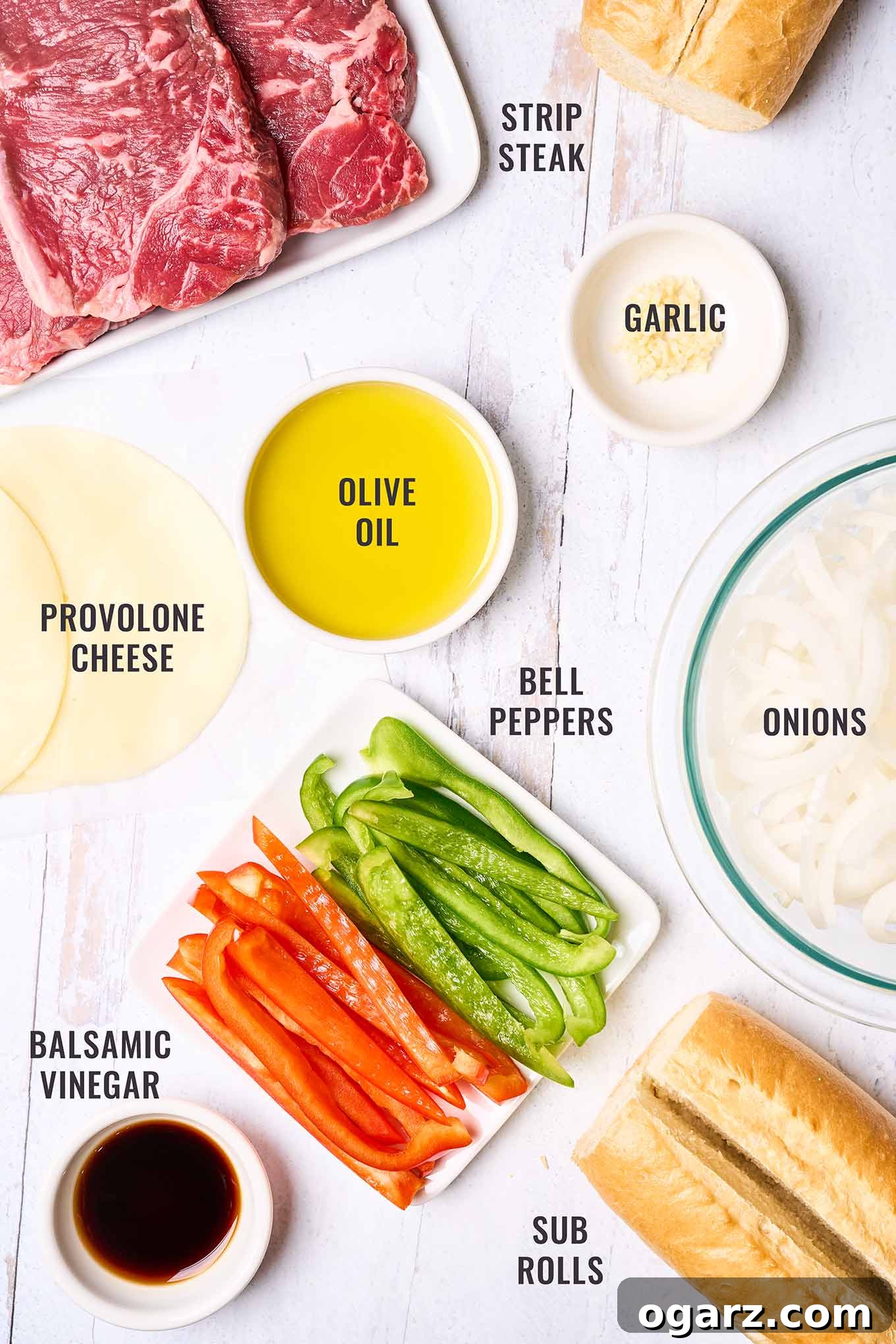 Philly's Signature Cheesesteak Indulgence 3 A labeled, overhead view of the ingredients needed to make philly cheesesteak sandwiches at home; strip steak, sub rolls, provolone cheese, olive oil, garlic, onions, red and green bell peppers, and balsamic vinegar.