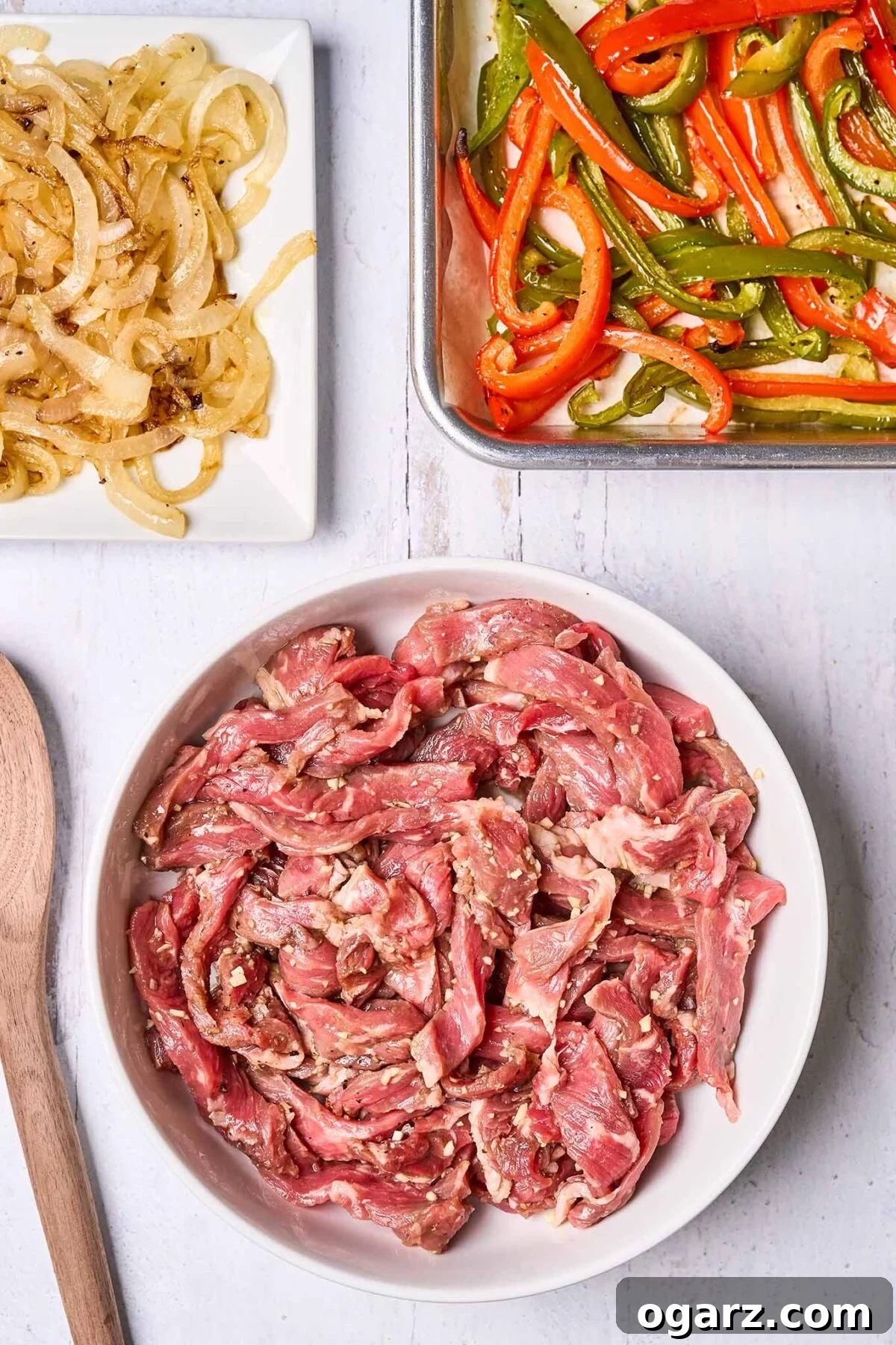 Philly's Signature Cheesesteak Indulgence 4 A parchment-lined sheet pan with strips of roasted red and green bell pepper, a plate filled with sautéed onions, and a bowl filled with marinated strips of steak, ready for pan-searing.