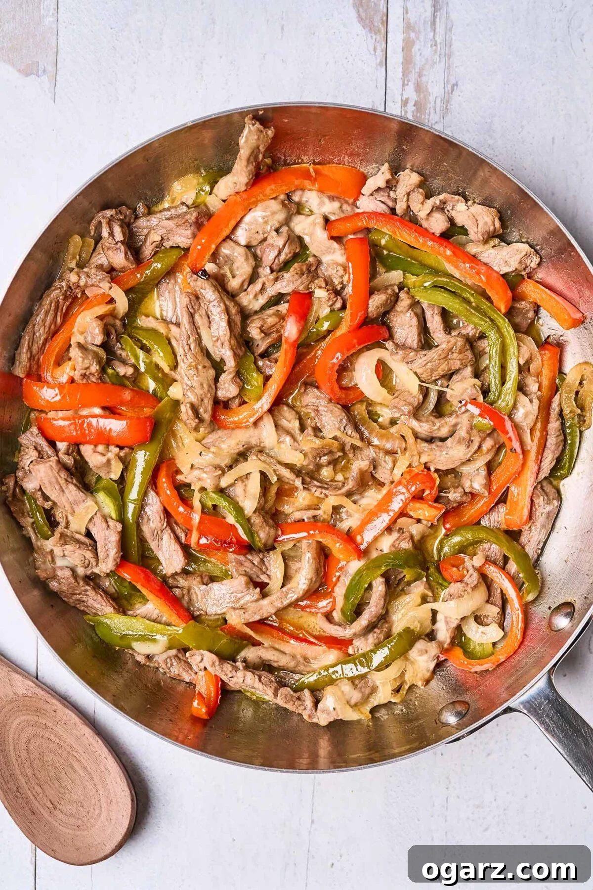 Philly's Signature Cheesesteak Indulgence 5 A frying pan filled with strips of pan-seared steak, roasted bell pepper, sautéed onions, and melted provolone cheese - ready to be spooned into sub rolls, a wooden spoon in the background.