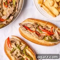 Two Philly cheesesteak sandwiches with peppers, onions, and provolone cheese on toasted sub rolls, a plate of steak fries and the frying pan with more steak, peppers, and onions in the background.