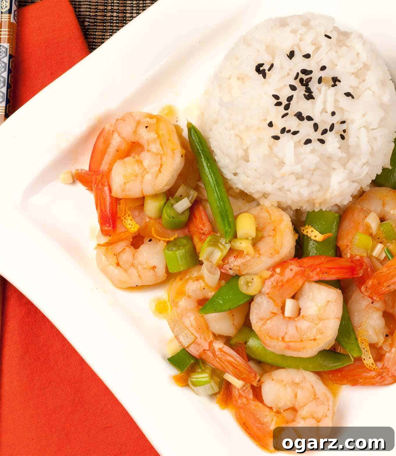 An overhead view of vibrant stir-fried shrimp with orange peel, crisp snap peas, and fresh scallions, served elegantly over fluffy white rice topped with black sesame seeds.