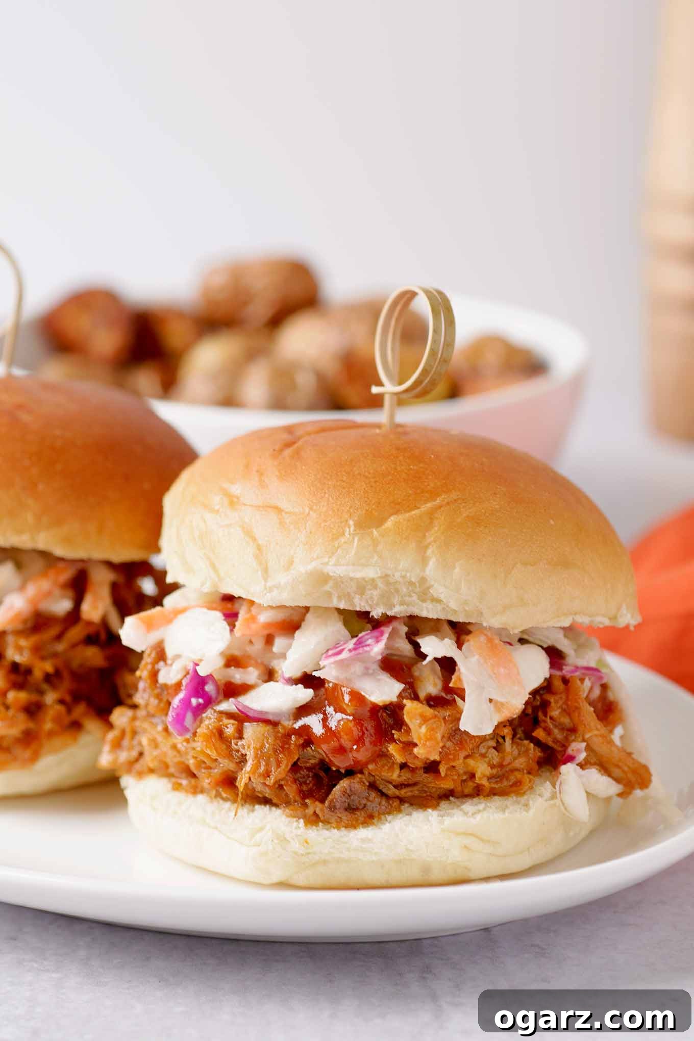 Perfectly Tender Oven-Roasted Pulled Pork 2 A pulled pork sandwich on a soft roll, topped with coleslaw and barbecue sauce, a bowl of roasted potatoes in the background.