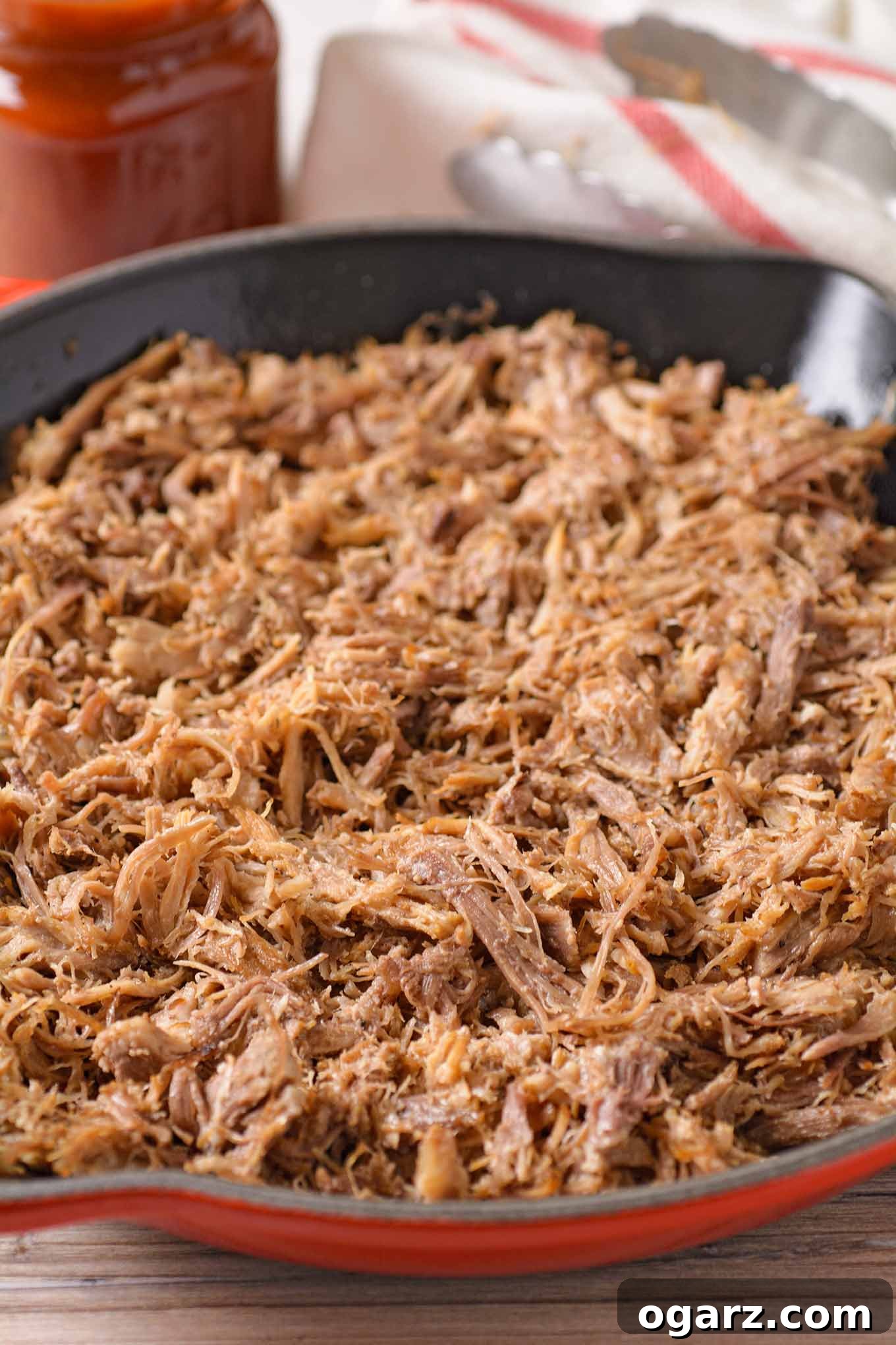 Perfectly Tender Oven-Roasted Pulled Pork 5 A close up of pulled pork in a cast iron skillet before adding sauce, a jar of sauce, kitchen towel, and tongs in the backgroud.
