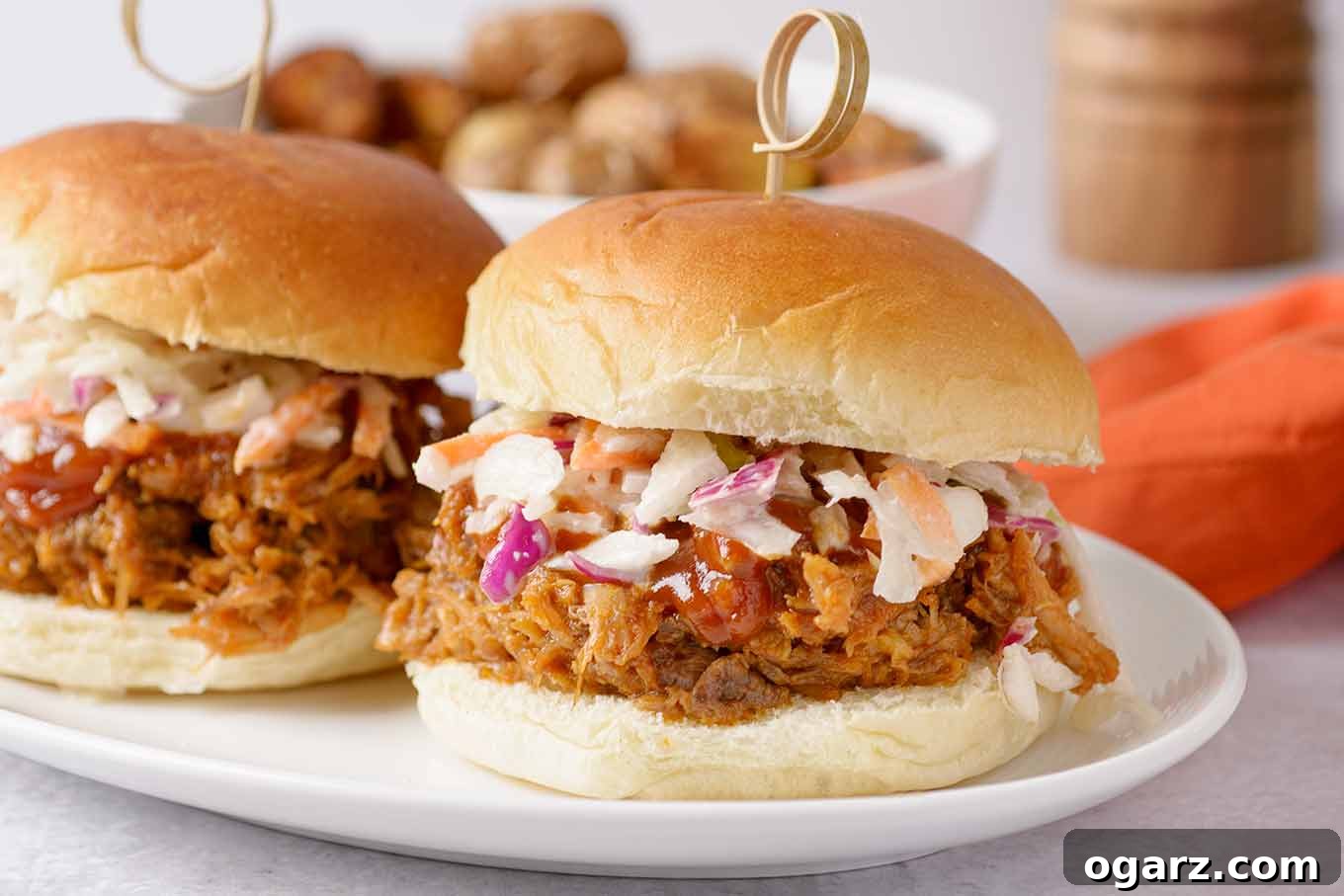 Perfectly Tender Oven-Roasted Pulled Pork 6 A close up of two pulled pork sandwiches on a plate, topped with extra barbecue sauce and homemade coleslaw, secured with a bamboo pick.