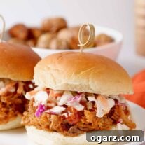 A pulled pork sandwich on a soft roll, topped with coleslaw and barbecue sauce, a bowl of roasted potatoes in the background.