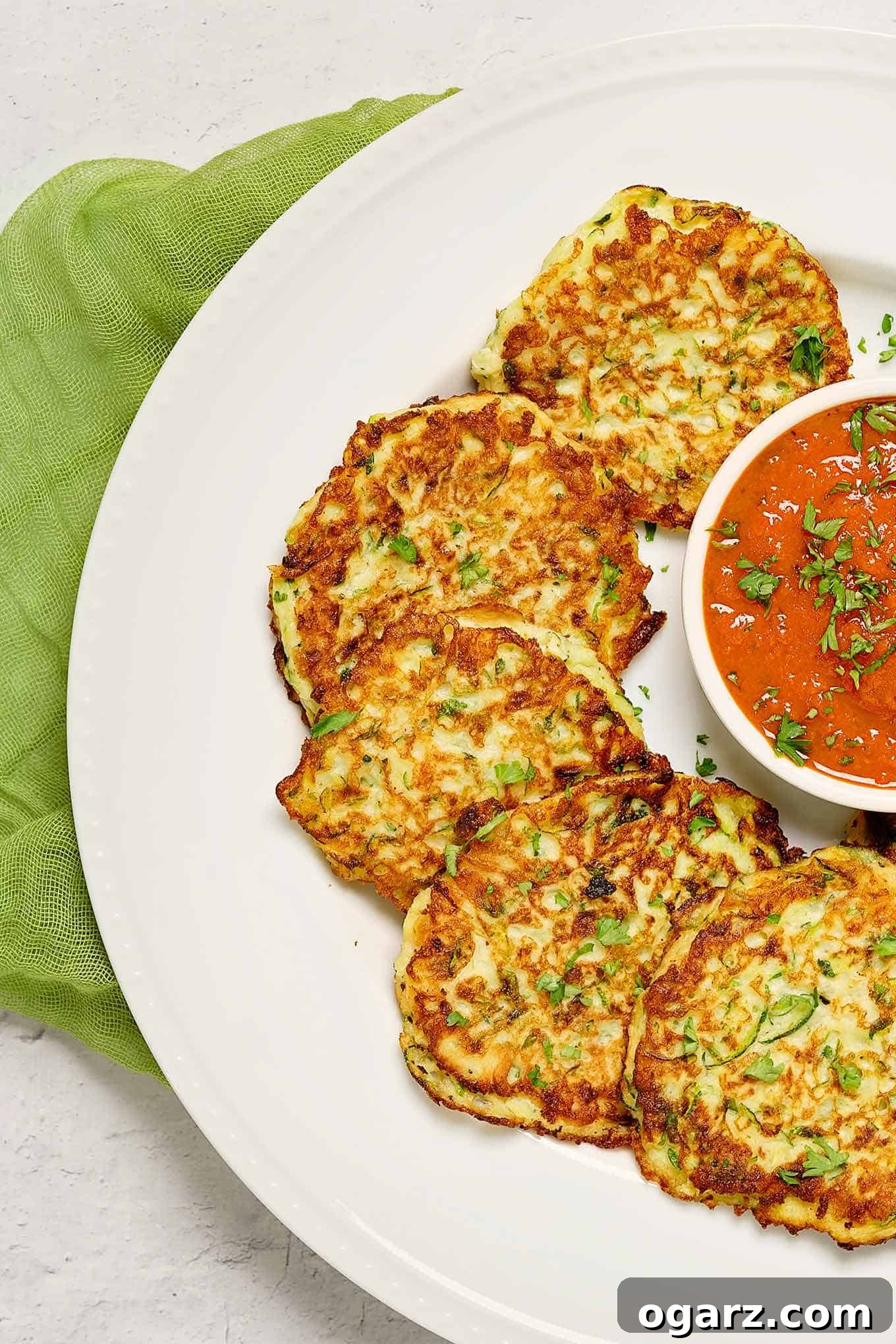 Crispy Parmesan Zucchini Bites 3 An overhead view of golden brown zucchini fritters on a round serving plate with marinara sauce, a green linen napkin under the plate.