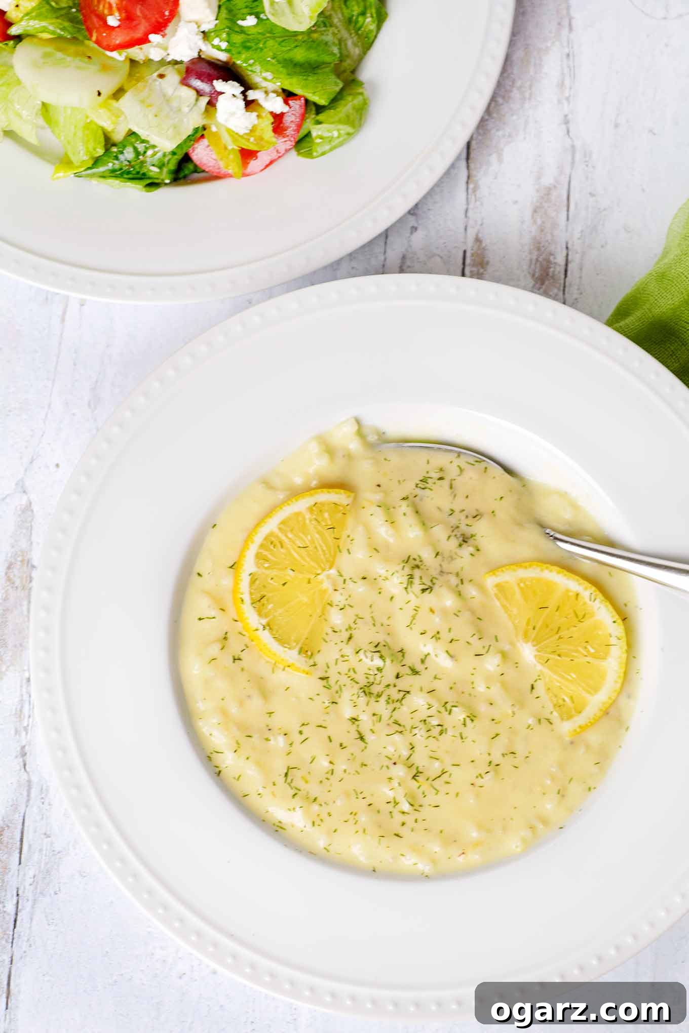 Avgolemono: Velvety Greek Lemon Soup 2 A vibrant white bowl of traditional Greek Avgolemono soup, elegantly garnished with fresh lemon slices and finely chopped dill. A crisp Greek salad sits invitingly on the side, highlighting a complete and fresh Mediterranean meal.