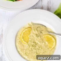 A white bowl of egg and lemon soup garnished with lemon wheels and fresh chopped dill with a Greek salad on the side.
