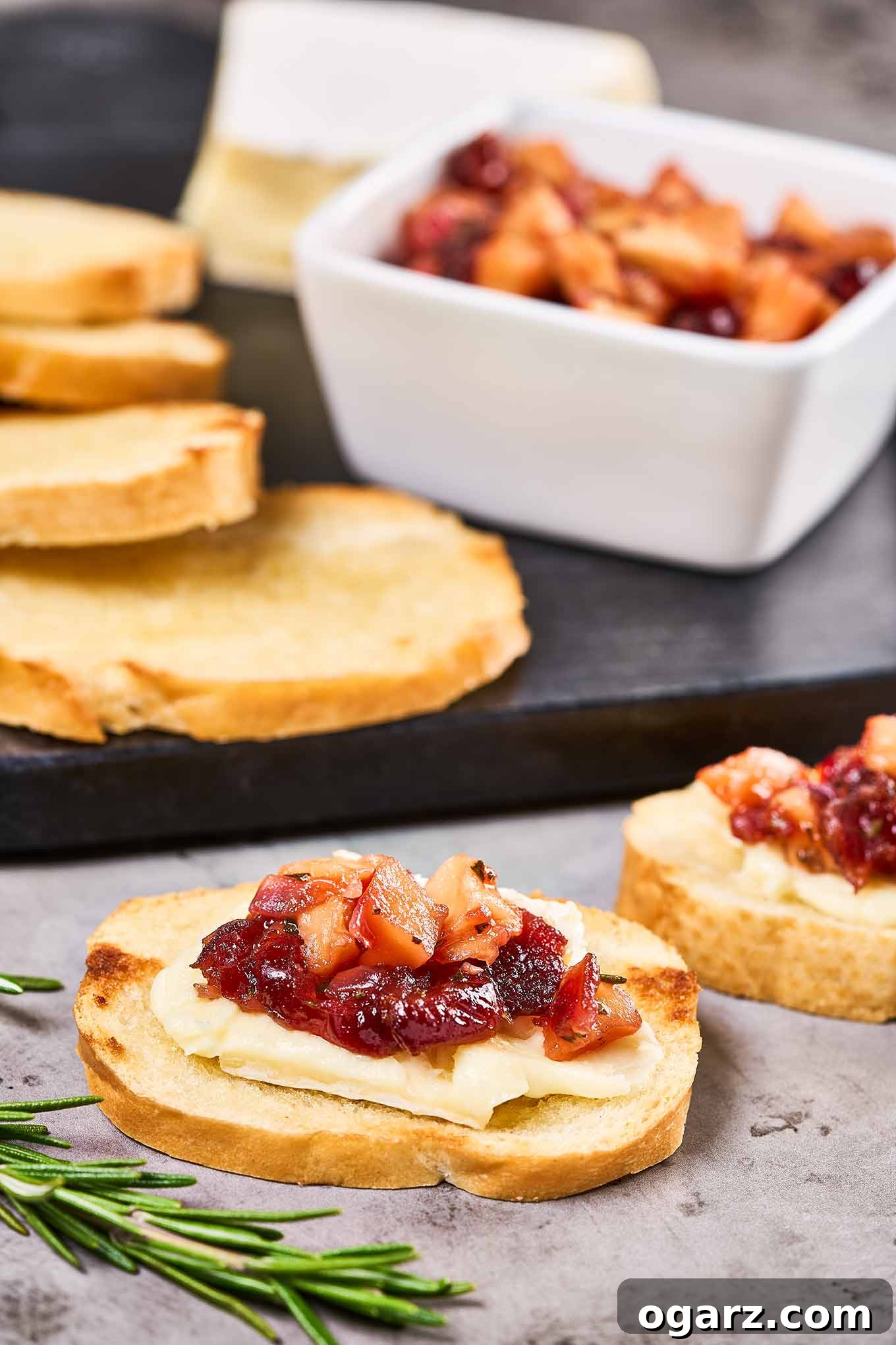 Two brie cheese and cranberry chutney crostini with a cheese board, more plain crostini, and a bowl of chutney in the background.