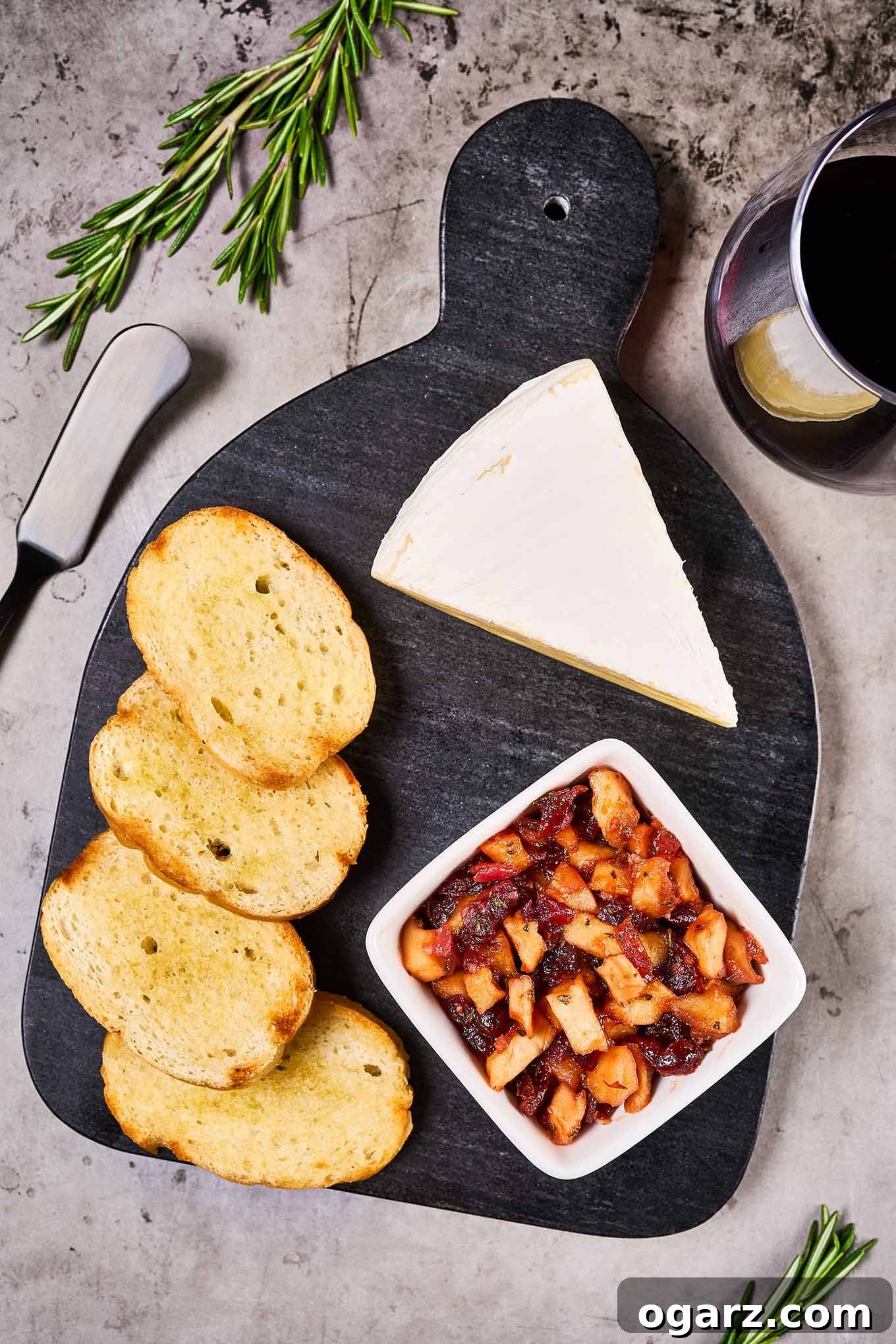 A wooden serving board with slices of toasted baguette, a dish of apple cranberry chutney, and a small wedge of Brie cheese, a glass of red wine to the side.
