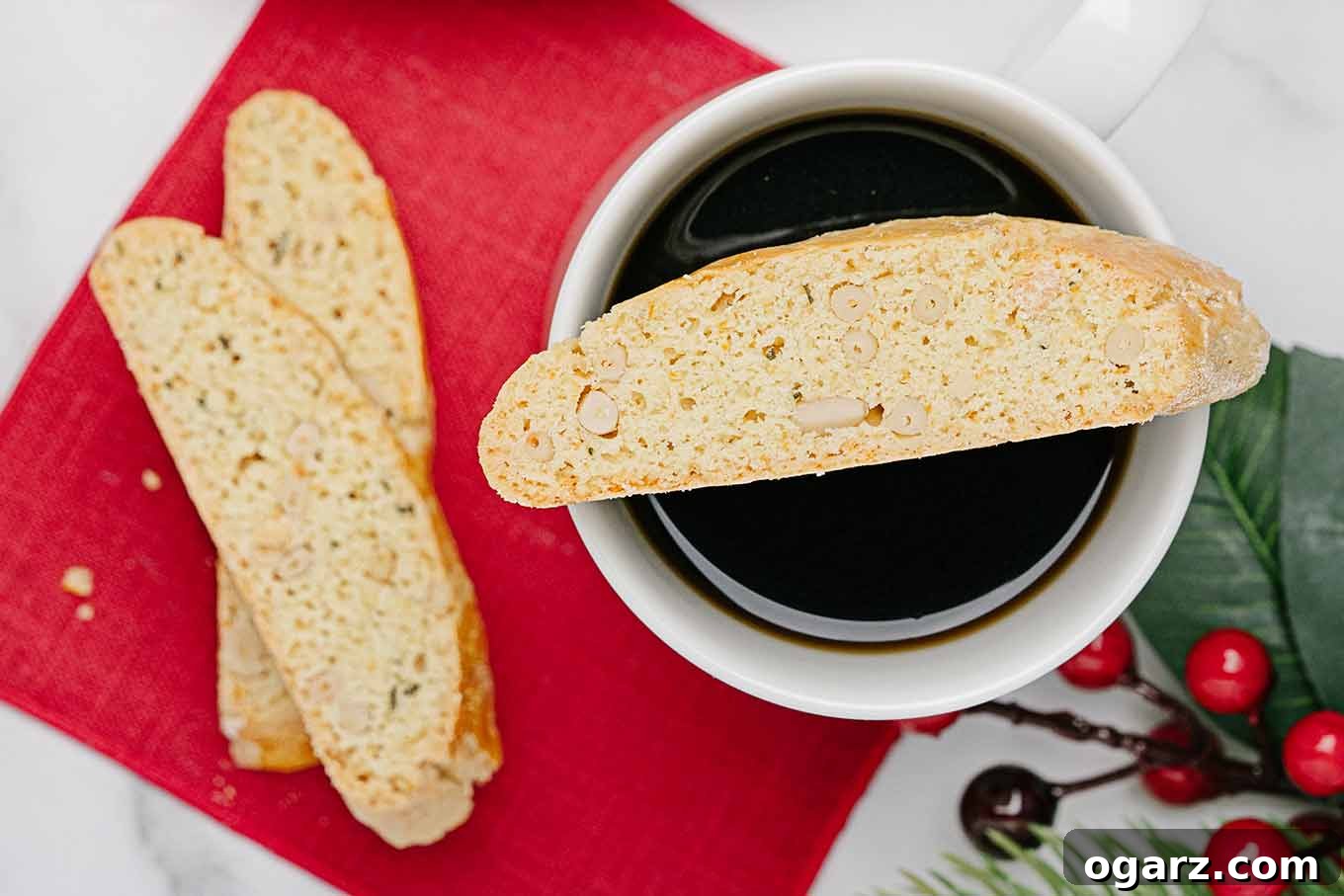 Golden Buttery Orange Biscotti 8 An overhead view of orange rosemary biscotti on top of a mug of coffee, a red napkin underneath, two more biscotti and a sprig of holly to the side.