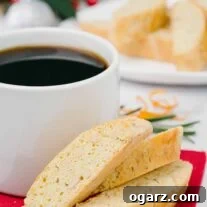 Buttery biscotti with orange, pine nuts, and rosemary on a red napkin next to a mug of coffee, more biscotti and Christmas table decor in the background.