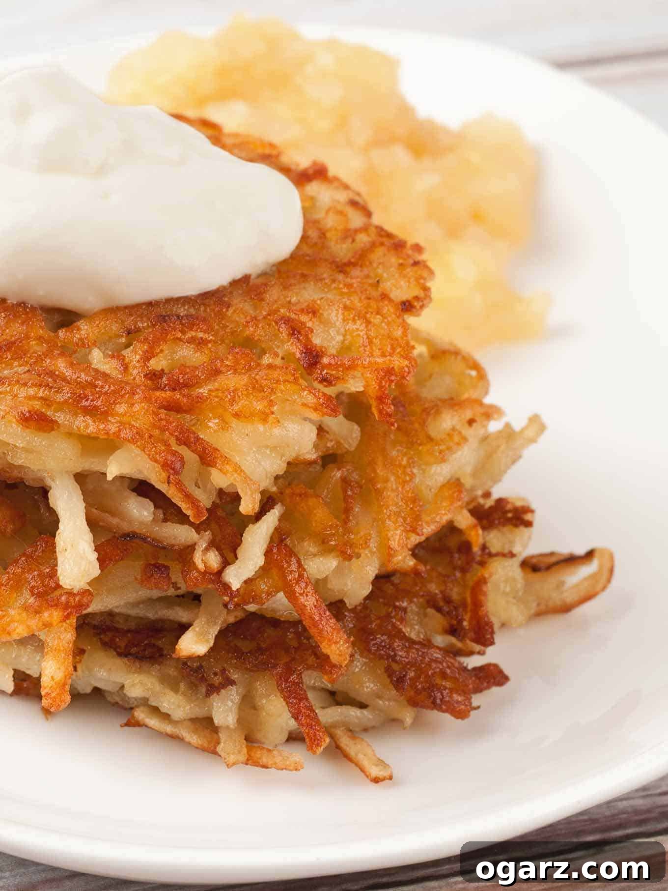 Golden Crispy Potato Latkes 2 Golden crispy potato latkes stacked on a white plate, topped with sour cream and served with applesauce.
