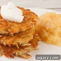 Golden crispy potato latkes stacked on a white plate, topped with sour cream and served with applesauce.