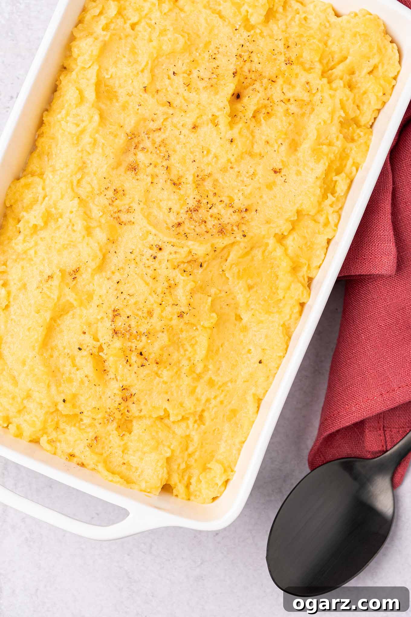 A white, oblong casserole dish of creamy mashed rutabagas sprinkled with nutmeg, a serving spoon and linen napkin on the side.