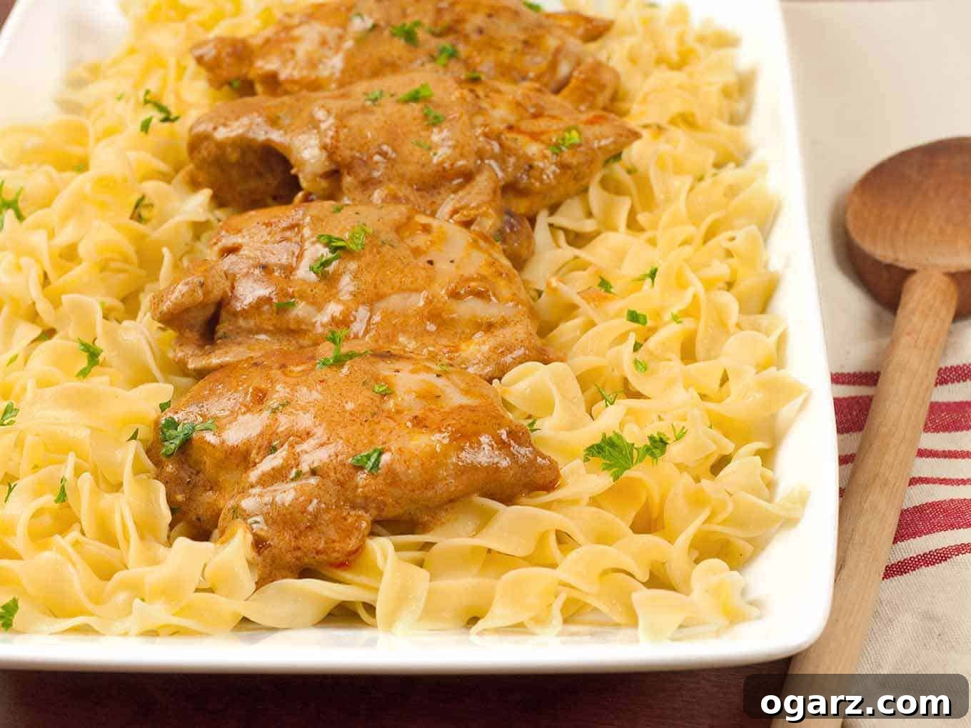 Easy Chicken Paprikash 2 Easy Chicken Paprikas served over a bed of buttered egg noodles, garnished with chopped parsley on a rectangular white platter.