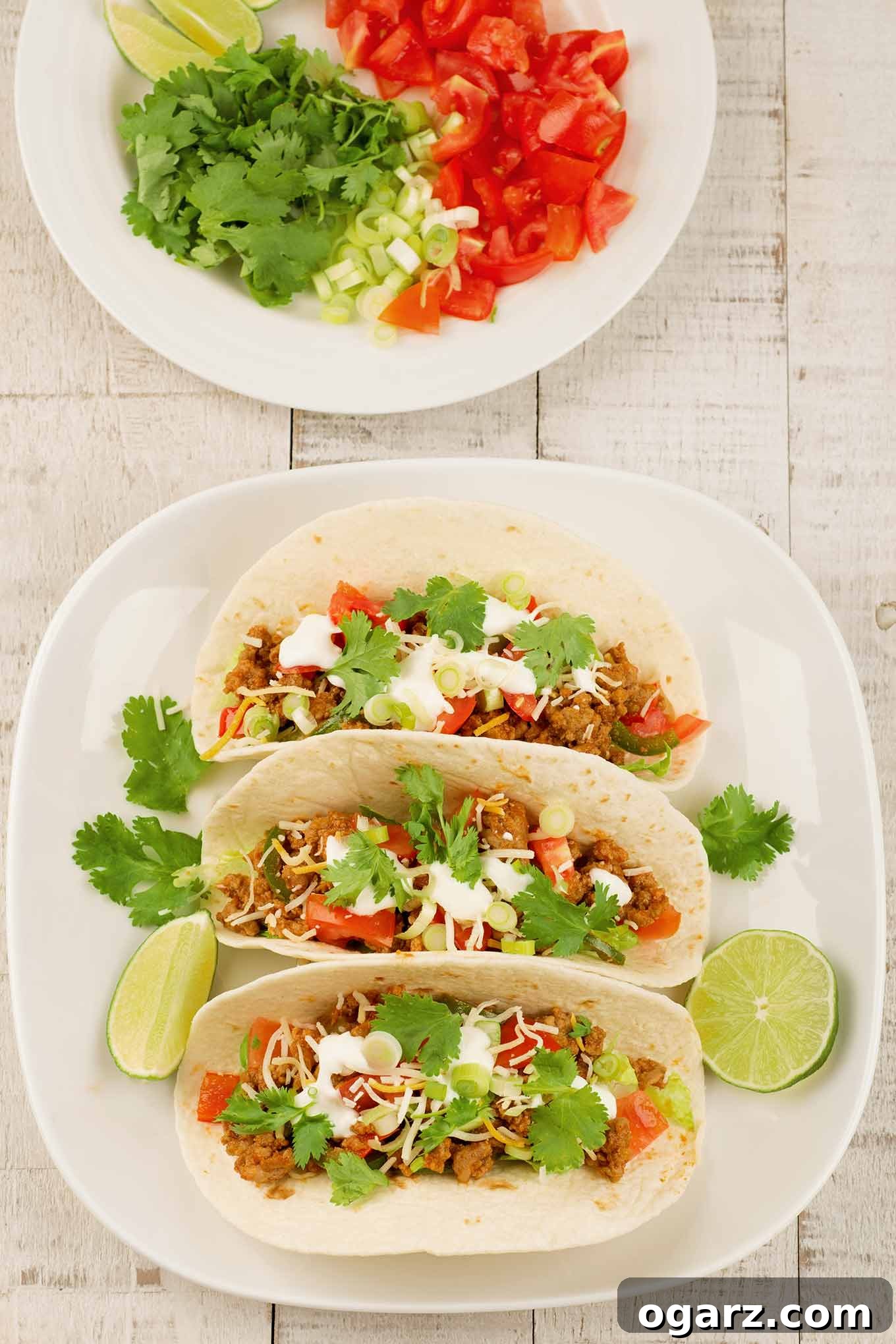 An overhead view of a plate of ground turkey tacos on a light wooden surface, next to a plate of toppings in the background.
