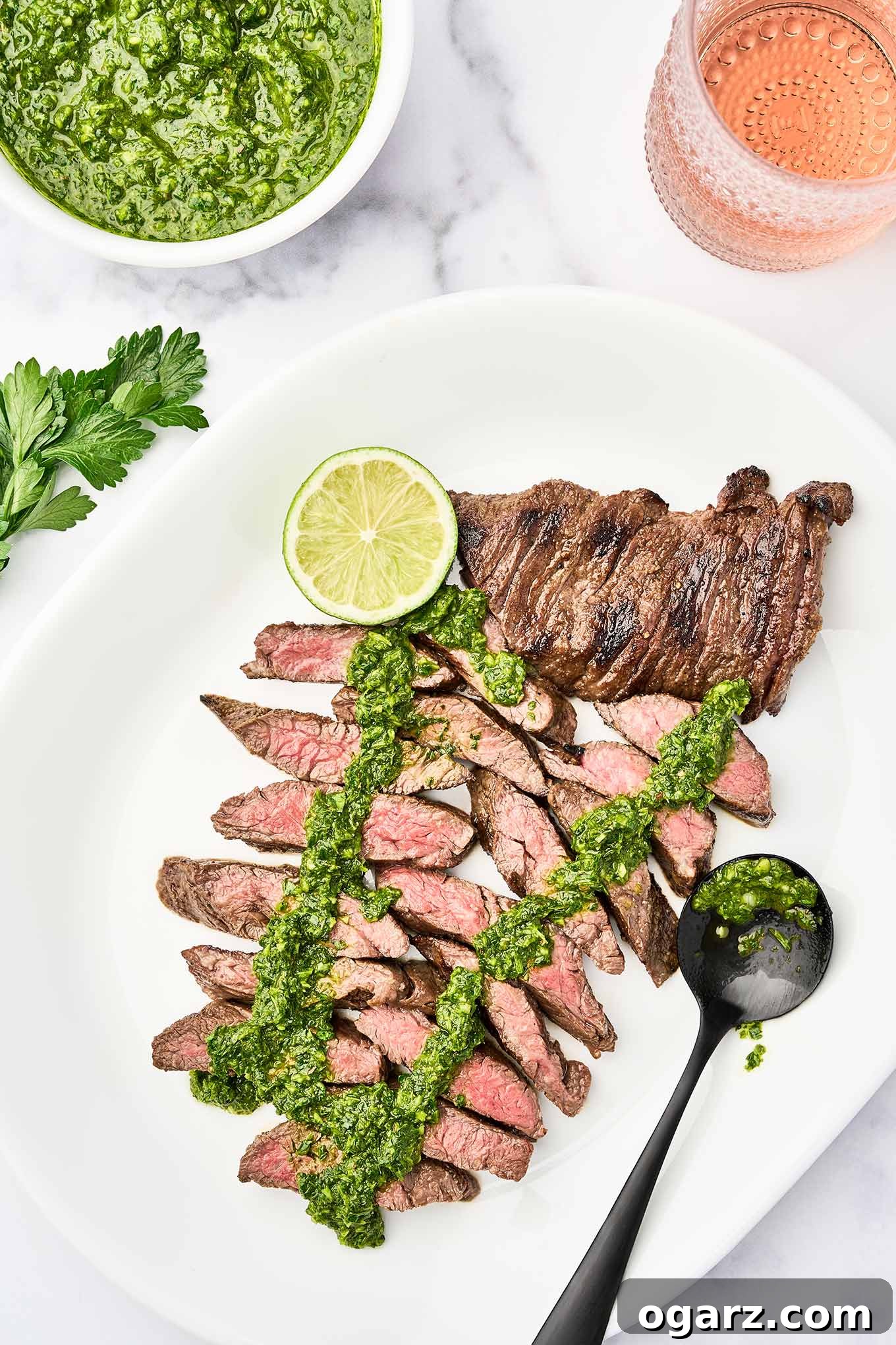A vibrant platter featuring perfectly grilled, medium-rare skirt steak, generously topped with fresh green chimichurri sauce and garnished with a lime wedge. A bowl of chimichurri and a glass of rosé are blurred in the background, suggesting a delightful meal.