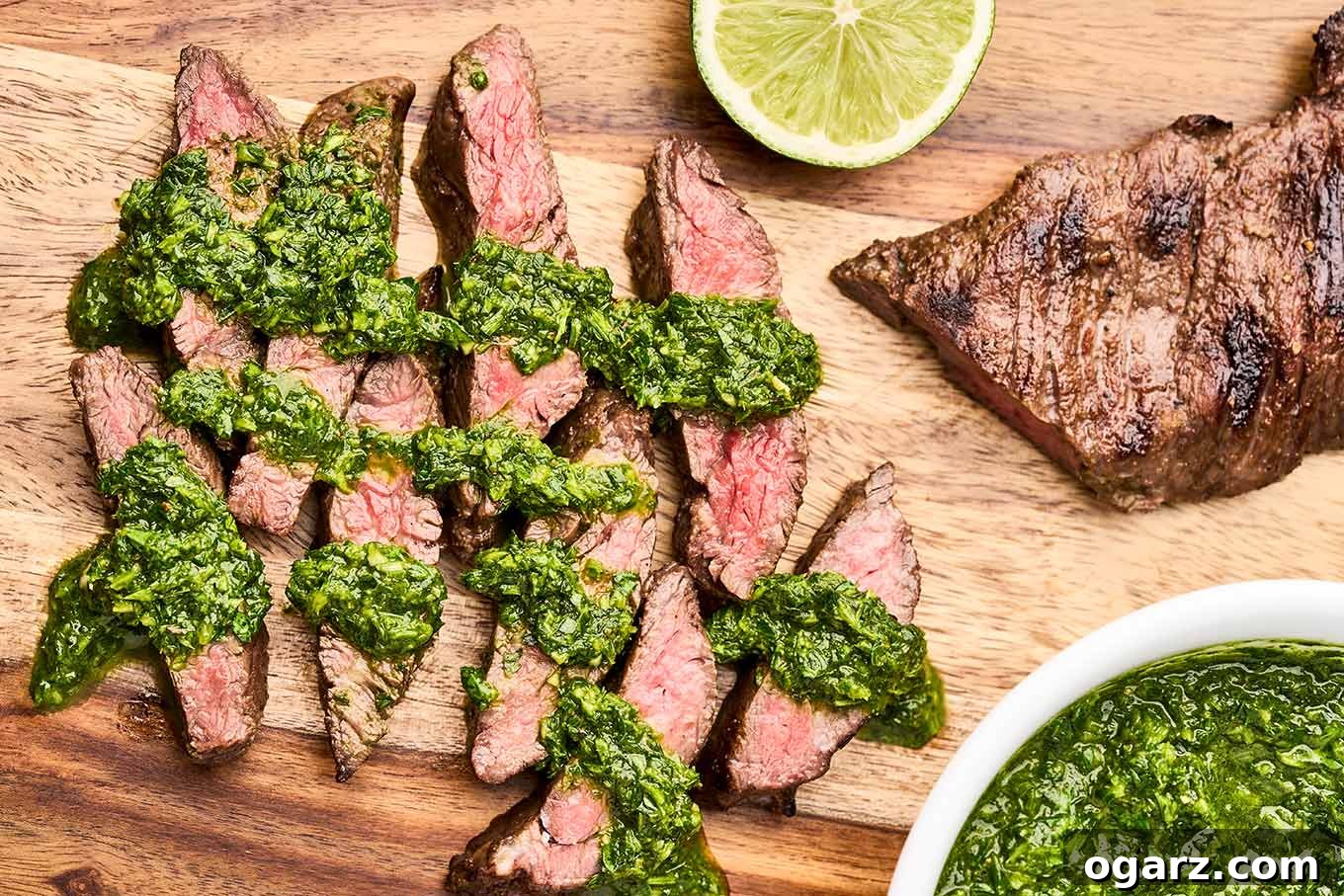 Close-up of thinly sliced grilled skirt steak, beautifully arranged on a wooden cutting board, with a generous drizzle of vibrant green chimichurri sauce and fresh lime wedges for garnish.
